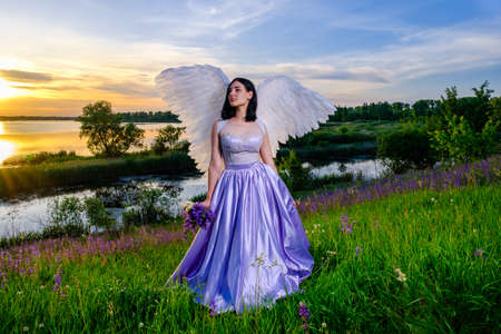 young beautiful white girl with angel wings stands on a hillside with violet wildflowers bouquet in her hands.の写真素材