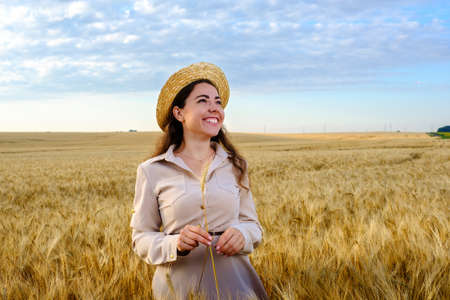 pretty young long-haired woman in a straw hat holds stalk of wheat in a field at sunrise and happy smiling. Copy spaceの写真素材