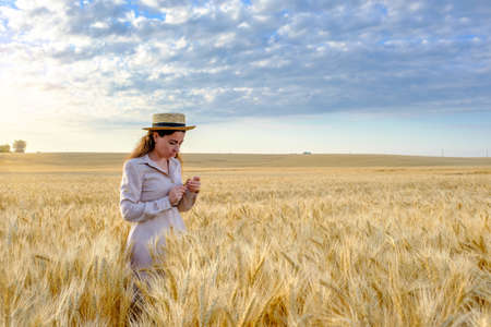 young long-haired woman agronomist in a straw hat holds stalk of wheat in a field at sunrise. Copy spaceの写真素材