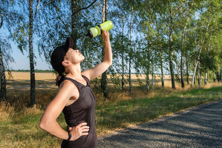 Resting young caucasian woman pouring water from a plastic bottle on her face after hard workout on the country road. Thirst, heat, restoration of strength and water balance in the body.の写真素材