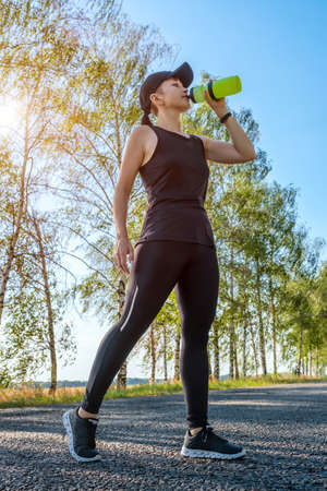Resting young caucasian woman drinks water from a plastic bottle after hard workout on the countryside road. Thirst, heat, restoration of strength and water balance in the body. Vertical orientationの写真素材