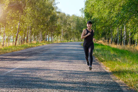 young white fitness woman runs along country road side, front view. Workout, fitness, marathon concept. Copy space for textの写真素材