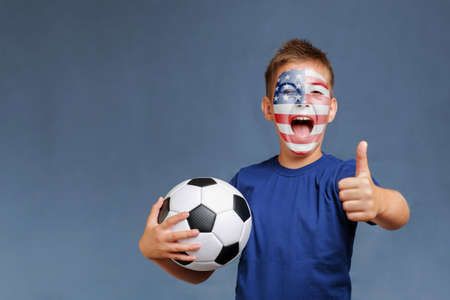 Handsome laughing American football fan holds soccer ball and gesturing thumbs up on gray background. Ukrainian flag on painted face. Copy space for text.の写真素材