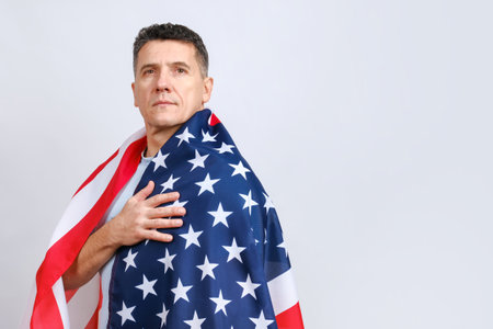 Handsome middle-aged man covered with American flag puts hand on his heart and looking at camera while standing against grey background. Copy space for textの写真素材
