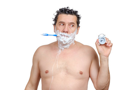 Sad middle-aged white man with toothbrush in his mouth, shave, washing head, looks on alarm clock in hand and being late for work or meeting or overslept isolated on white backgroundの写真素材