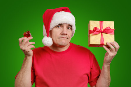 Disappointment middle-aged man in Santa hat and dissatisfied face holds small and big Christmas gifts on green backgroundの写真素材