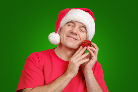 Satisfied middle-aged caucasian man in Santa hat receive small Christmas gift, holds near his face and smiling with closed eyes on green festive background. Copy space for text in right sideの写真素材