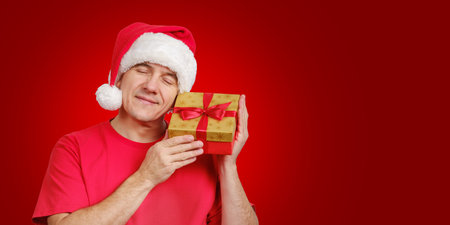 Middle-aged caucasian man in Santa hat receive Christmas gift, holds near his face and smiling with closed eyes on red festive background. Copy space for text in right sideの写真素材
