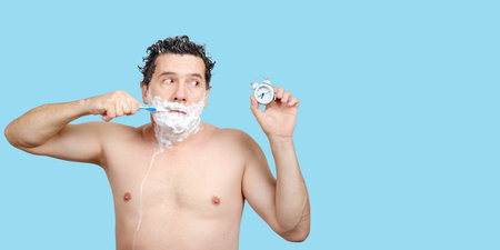 Sad middle-aged white man holds toothbrush in his mouth, shave, washing head, looks on alarm clock in hand and being late for work or meeting or overslept on blue background, copy space for textの写真素材