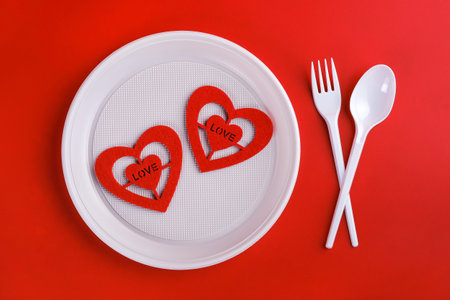 Top view of plastic white disposable fork, spoon, plate and red hearts isolated on red background, valentines day for poor people conceptの写真素材