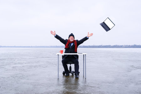 Senior smiling businessman in suit sits with outstretched hands at a table in the middle of a frozen lake in winter and throws away his laptop. Copy space for text on screen of deviceの写真素材