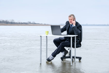 elderly bearded smiling businessman in suit and skates, smiling while talking on smartphone, works with laptop on table in the middle of a frozen lake. Copy space for text, work on vacation conceptの写真素材