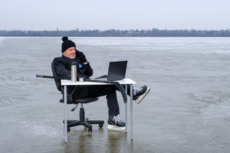 elderly bearded smiling businessman in suit and skates holds hockey stick, smiling while talking on smartphone, works with laptop on table in the middle of a frozen lake. Copy space for textの写真素材