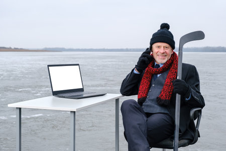 elderly bearded smiling businessman in suit holds hockey stick, smiling while talking on smartphone, works with laptop on table in the middle of a frozen lake. Copy space for text on device screenの写真素材