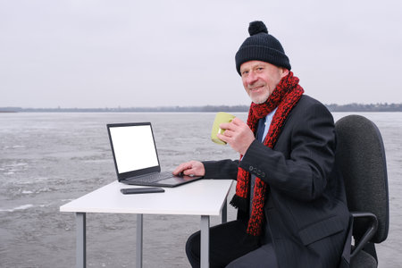 elderly bearded smiling businessman in suit looking to the camera, drinks coffee and works with laptop on table in the middle of a frozen lake. Copy space for text on device screenの写真素材