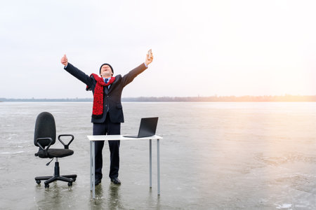 Senior smiling bearded businessman in suit stands with outstretched hands and gesturing thumbs up near table and laptop in the middle of a frozen lake in winter. Copy space for text, good deal conceptの写真素材