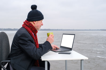 elderly bearded smiling businessman in suit looking to the camera, drinks coffee and works with laptop on table in the middle of a frozen lake. Profile view, copy space for text on device screenの写真素材