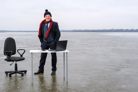 Senior smiling bearded businessman in suit stands near table with laptop and chair in the middle of a frozen lake in winter. Copy space for text, work and leisure conceptの写真素材