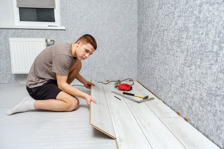 Young male repairman is laying wooden panel of laminate floor indoors on the knees. Laminate flooring, copy space.の写真素材