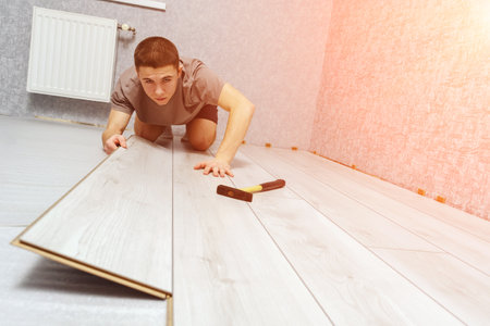 Young male repairman is laying wooden panel of laminate floor indoors. Laminate flooring, copy space. Selective focusの写真素材