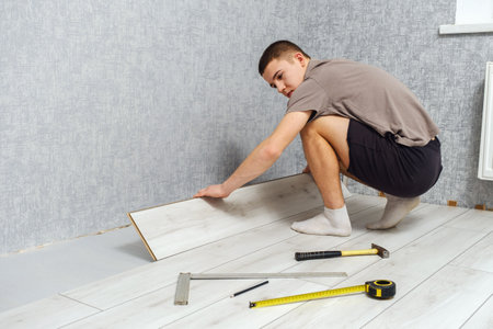 Young male repairman is laying wooden panel of laminate floor indoors on the knees. Laminate flooring, copy space, back viewの写真素材