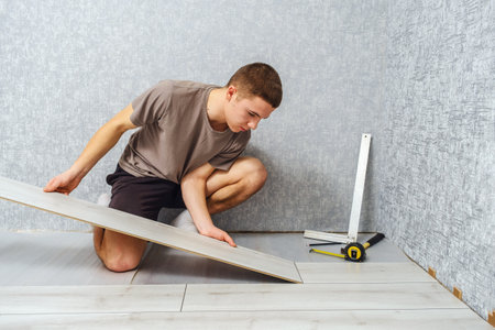 Young male repairman is laying wooden panel of laminate floor indoors on the knees. Laminate flooringの写真素材