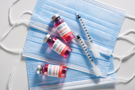Three glass vaccine vials and syringes for vaccination against the COVID-19, SARS-CoV-2 coronavirus pandemic, lying on surgical protective masks, flat layの写真素材