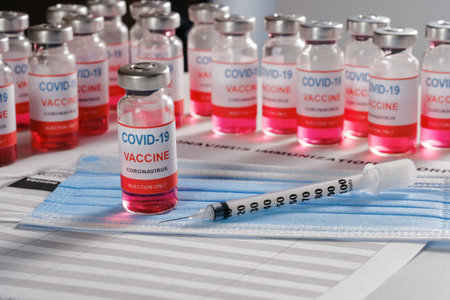 Vaccine vial, syringe and many glass bottles on background for vaccination against COVID-19 SARS-CoV-2 coronavirus pandemic lying on surgical mask and immunization document.の写真素材