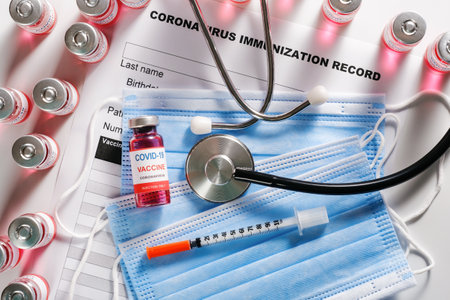 Stethoscope, syringe and vaccine vial glass bottles for vaccination against COVID-19 SARS-CoV-2 coronavirus pandemic lying on surgical masks and on the immunization document. Top view, flat layの写真素材