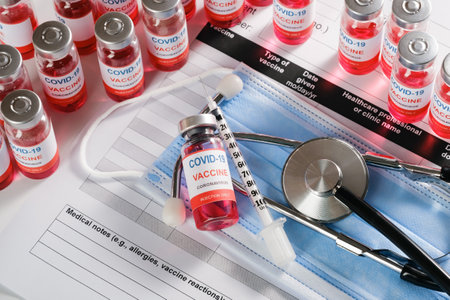 Stethoscope, syringe and vaccine vial glass bottles for vaccination against COVID-19 SARS-CoV-2 coronavirus pandemic lying on surgical masks and on the immunization document.の写真素材