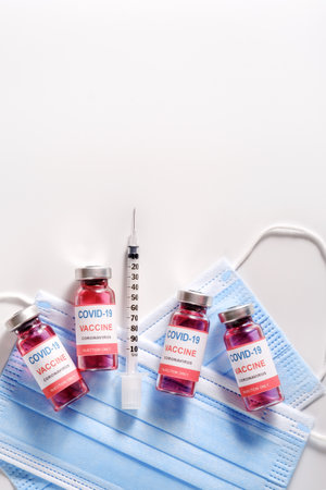 Glass vaccine vials and syringe for vaccination against the COVID-19, SARS-CoV-2 coronavirus pandemic, lying on surgical protective masks, flat lay, copy space, vertical orientationの写真素材