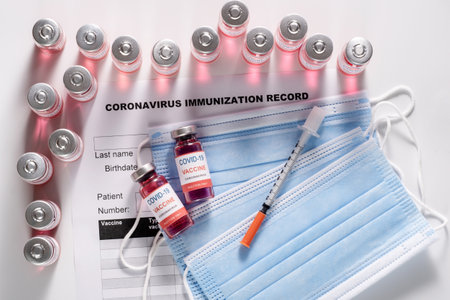 Vaccine glass vials and syringe for vaccination against COVID-19 SARS-CoV-2 coronavirus pandemic lying on the surgical protective mask and on the immunization document, flat layの写真素材