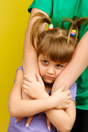 Portrait of sad little caucasian girl with pigtails and bruised eye looking for protection in her mothers arms on yellow background. Vertical orientationの写真素材