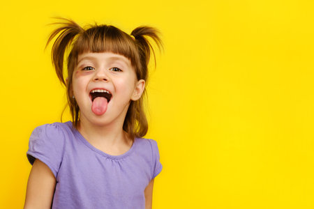 Close-up of laughing funny little bad girl with bruised eye showing her tongue, looking at camera on yellow background.の写真素材