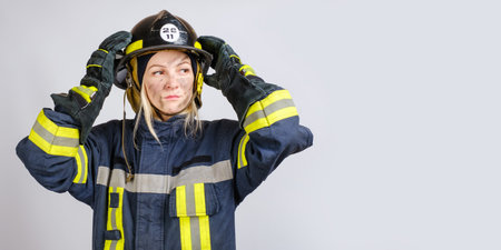 young brave woman in uniform of firefighter puts hardhat on her head and looking away on gray background, copy space for textの写真素材