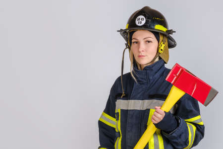 Young brave woman in uniform and hardhat of firefighter with axe in hands looking at camera on gray background, copy spaceの写真素材