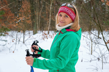 Elderly woman standing with nordic walking poles in winter forest, resting after exercise and looking at camera outdoors with smile.の写真素材