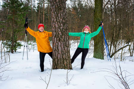 Smiling senior couple resting after walking with nordic walking poles and standing behind tree with outstretched arms in snowy winter forest. Active lifestyle concept.の写真素材