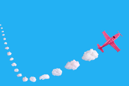 Pink airplane takes into a sharp turn on blue background with trail of abstract shapes of cloud....