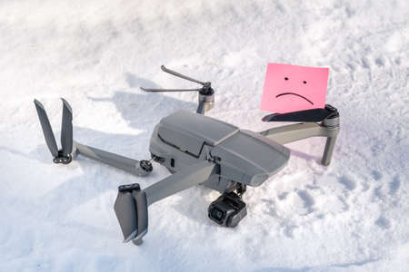 Sticky note with sorry smile in propeller blades. Broken gimbal camera and drone motor arm after crash on snow in winter outdoor closeup. Expensive repairs conceptの写真素材
