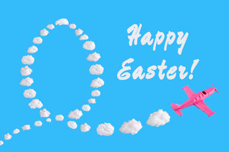 Pink airplane goes into a turn on blue background with text happy easter and easter egg of abstract shapes of cloud. Festive minimal composition, holiday conceptの写真素材