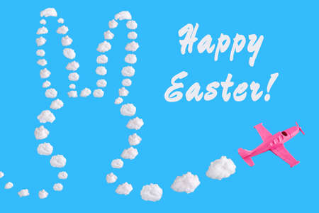Pink airplane goes into a turn on blue background with text happy easter and easter bunny of abstract shapes of cloud. Festive minimal composition, holiday conceptの写真素材