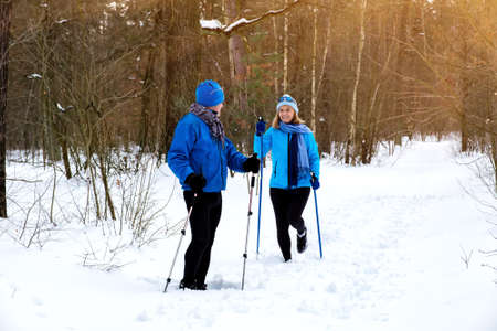 Man waiting woman on walking with nordic walking poles in snowy winter park. Elderly wife and husband doing healthy exercise outdoors. Active lifestyle after retirement concept.の写真素材