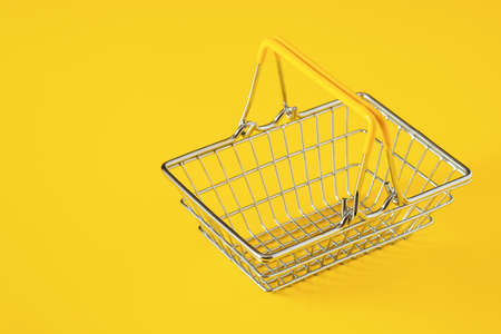 Empty metal wire shopping basket with yellow handles isolated on yellow background. Consumerism concept, copy space for text, close-up view, selective focusの写真素材