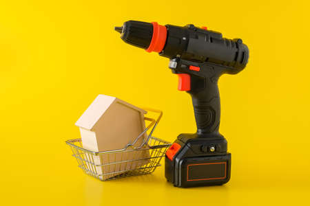 Modern black cordless screwdriver, drill with rechargeable battery and wooden house in shopping basket on yellow background, copy space for text, construction or renovation conceptの写真素材