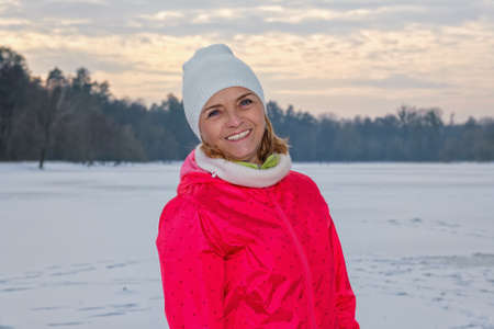 Elderly woman in white knitted hat standing in winter forest, resting after exercise and looking at camera outdoors with smile.の写真素材