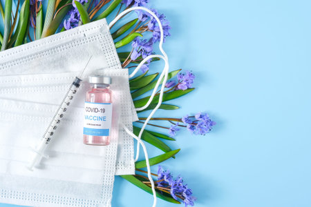 Vaccine, syringe, medical masks and snowdrop flowers on blue pastel background closeup.