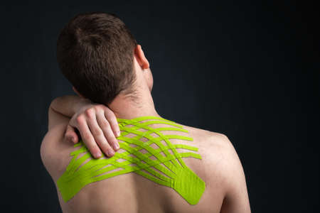 Hand of young man holding his neck with striped kinesiology medical tape applied to relieve back pain on dark background, back viewの写真素材