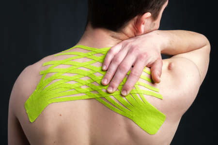 Hand of young man holding his neck with striped kinesiology medical tape applied to relieve back pain on dark background. Close-up, back viewの写真素材