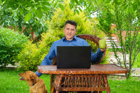 Mature Caucasian Man petting a dog, drinking coffee, analyzing web information and reading news via laptop in the backyard garden. Remote business conceptの写真素材